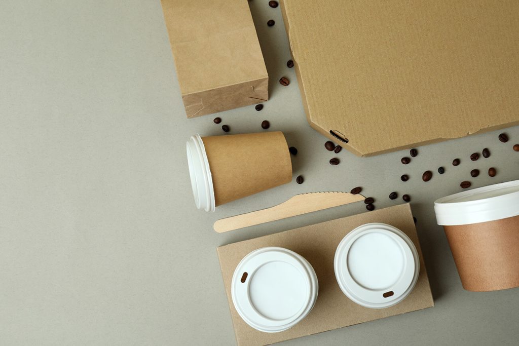 Assorted eco-friendly coffee takeout containers and cups with white lids, a cardboard box, a wooden knife, and scattered coffee beans on a neutral background.